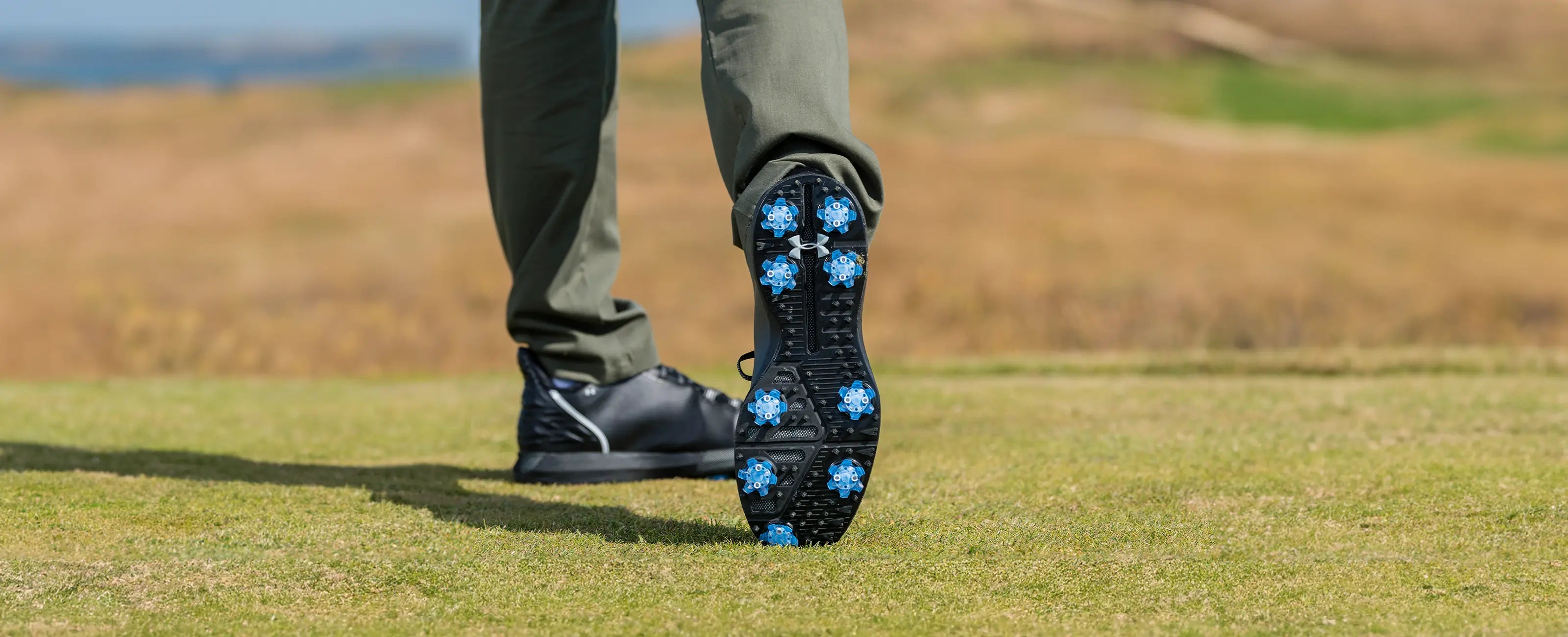 A person golfing wearing under armor shoes with blue Pivix golf spikes attached.
