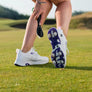 A golfer is adjusting their footwear, which is a white and purple golf shoe with cleats, while holding a shoe spike brush tool on a grassy course.