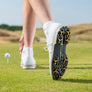A close-up of a golfer's foot in a white shoe with scorpion stinger golf spikes, standing on green grass near a golf ball on a tee.