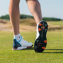 A person's leg is seen from behind, wearing a white golf shoe with silver tornado golf spikes and standing on grass.