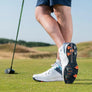 A close-up of a golfer's foot wearing a white golf shoe and silver tornado golf spikes on a grassy golf course, with a golf club visible in the background.
