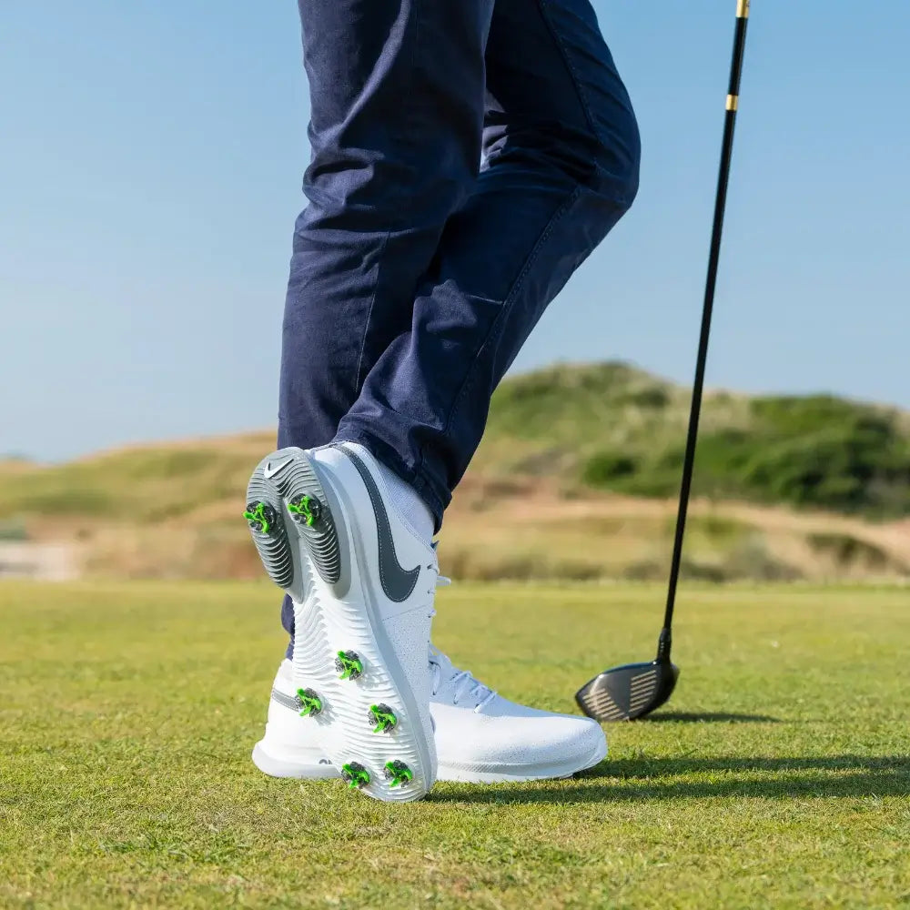 A person wearing white golf shoes with green tour flex pro spikes stands on a golf course, holding a golf club.