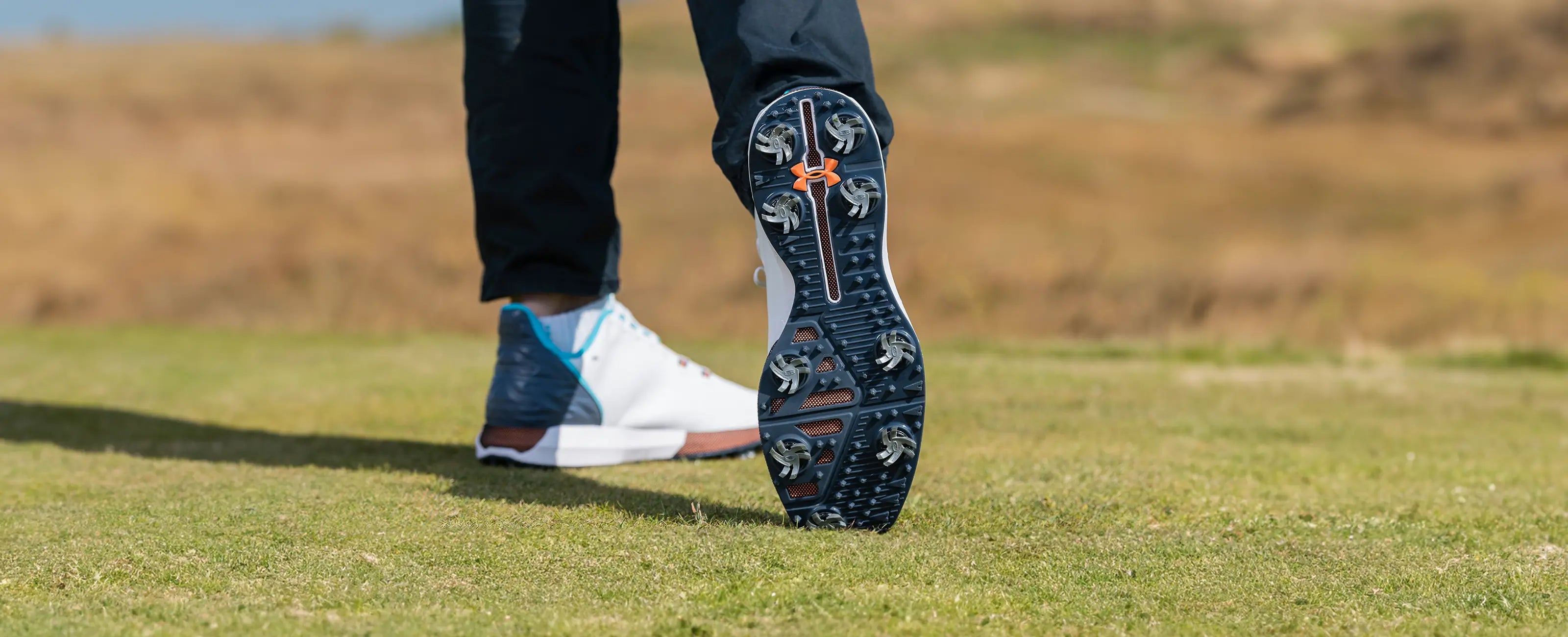 A person golfing while wearing under armor shoes with Silver Tornado spikes attached to them. 