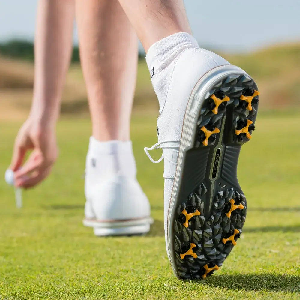 A close-up of a golfer's foot in a white shoe with scorpion stinger golf spikes, standing on green grass.