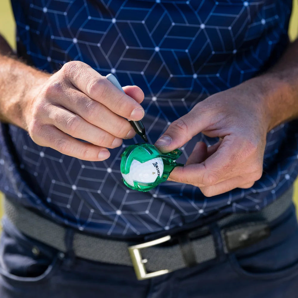 A person holds a golf ball with a green alignment tool in one hand and a black marker in the other, preparing to mark the ball.