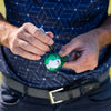 A person holds a golf ball with a green alignment tool in one hand and a black marker in the other, preparing to mark the ball.