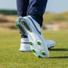 A close-up of a foot wearing a white shoe with green tour flex pro spikes, standing on grassy terrain.