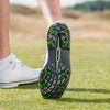A close-up of a foot wearing a white shoe with green tour flex pro spikes, standing on grassy terrain.
